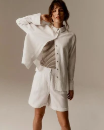 Model wearing a white button-down shirt over a striped top and white shorts against a plain studio background