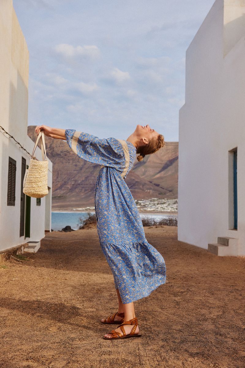 A blonde woman wearing a long blue floral dress, leather sandals, and holding a straw bag poses outdoors with white buildings.