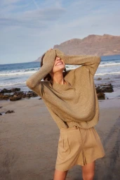 Model wearing a beige knit sweater and shorts covering her eyes on a sandy beach with the ocean and mountains behind her.
