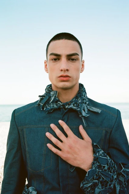 Close-up of a young adult man wearing a dark denim jacket with a light blue sky and horizon in the background on a sunny day.