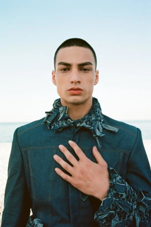 Close-up of a young adult man wearing a dark denim jacket with a light blue sky and horizon in the background on a sunny day.