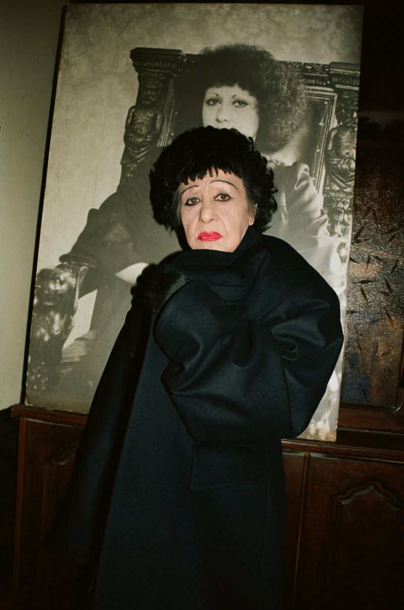 Portrait of an older woman in a black coat and wig, standing indoors in front of a large framed portrait, moody lighting.