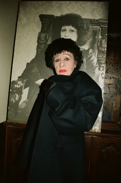 Portrait of an older woman in a black coat and wig, standing indoors in front of a large framed portrait, moody lighting.