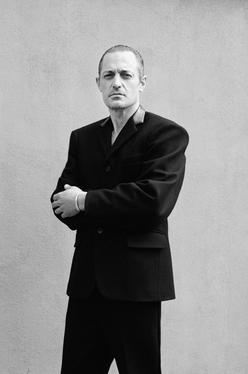 Black and white portrait of adult male in black suit with arms crossed against a light neutral background in an indoor setting.