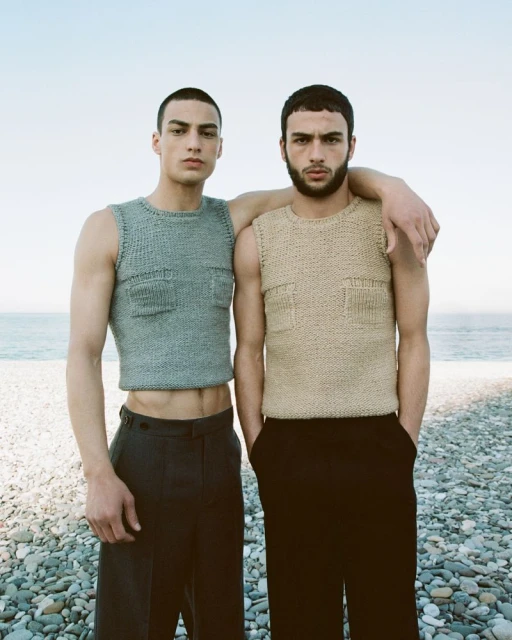 Two adult men pose on a rocky beach wearing knitted tank tops and black trousers against a backdrop of sea and sky.