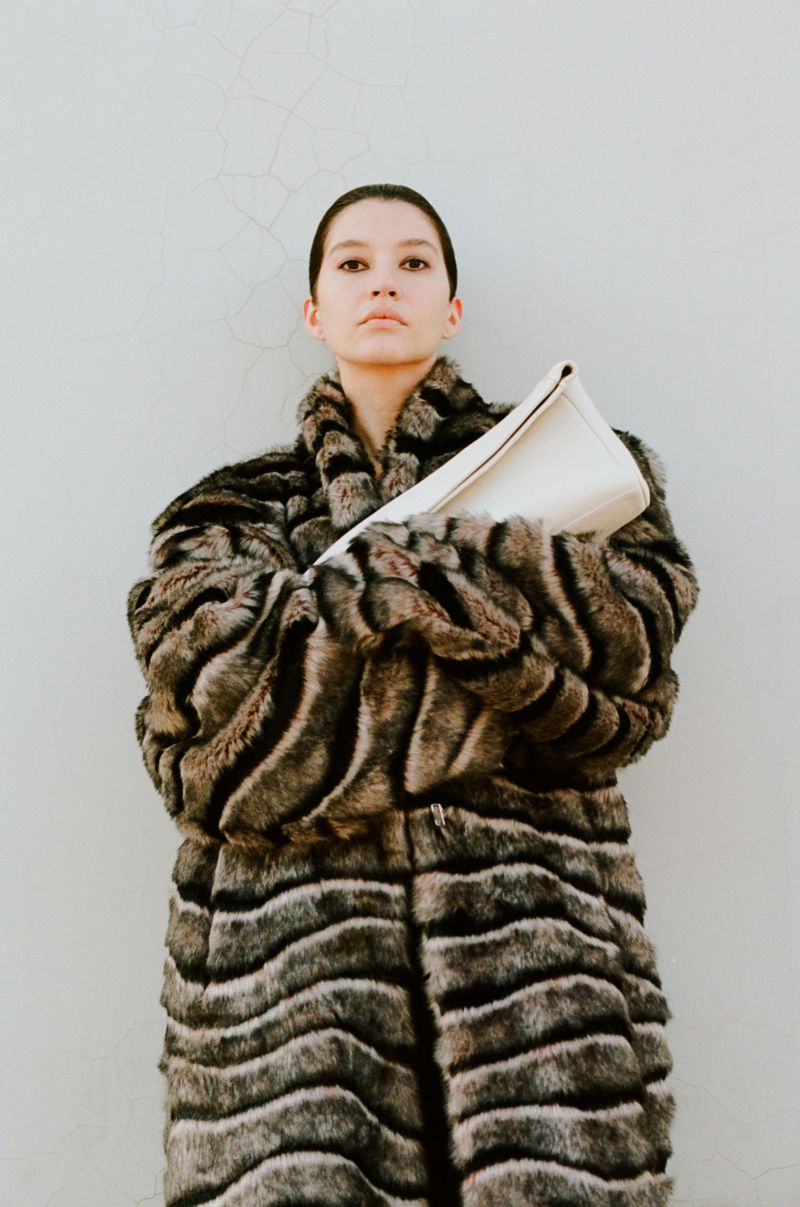 Model wearing an oversized striped faux fur coat while holding a white leather clutch bag against a pale wall.