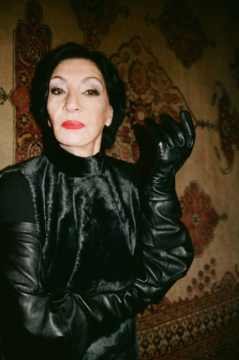 Portrait of a stylish adult woman wearing black faux fur top and leather gloves posing in front of a patterned rug indoors.
