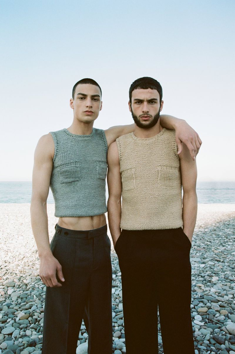 Two adult men wearing knitted tank tops and dark pants pose on a rocky beach with the ocean and clear sky in the background.