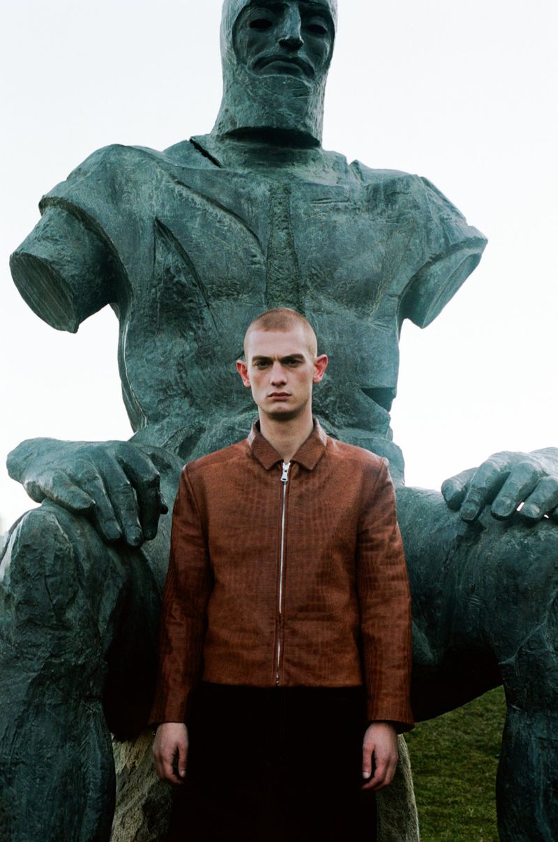 Adult male in a jacket stands in front of a large, weathered statue in an outdoor setting with muted lighting, creating an editorial feel.