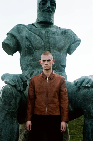 Adult male in a jacket stands in front of a large, weathered statue in an outdoor setting with muted lighting, creating an editorial feel.