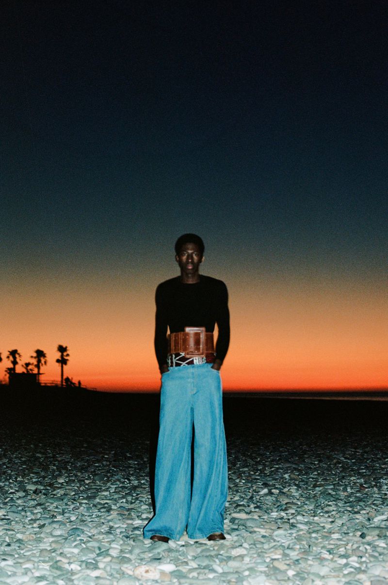 A person with dark skin is standing on a rocky beach wearing a black long-sleeve shirt and wide-leg jeans in front of an orange sunset.
