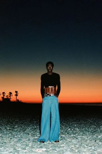 A person with dark skin is standing on a rocky beach wearing a black long-sleeve shirt and wide-leg jeans in front of an orange sunset.