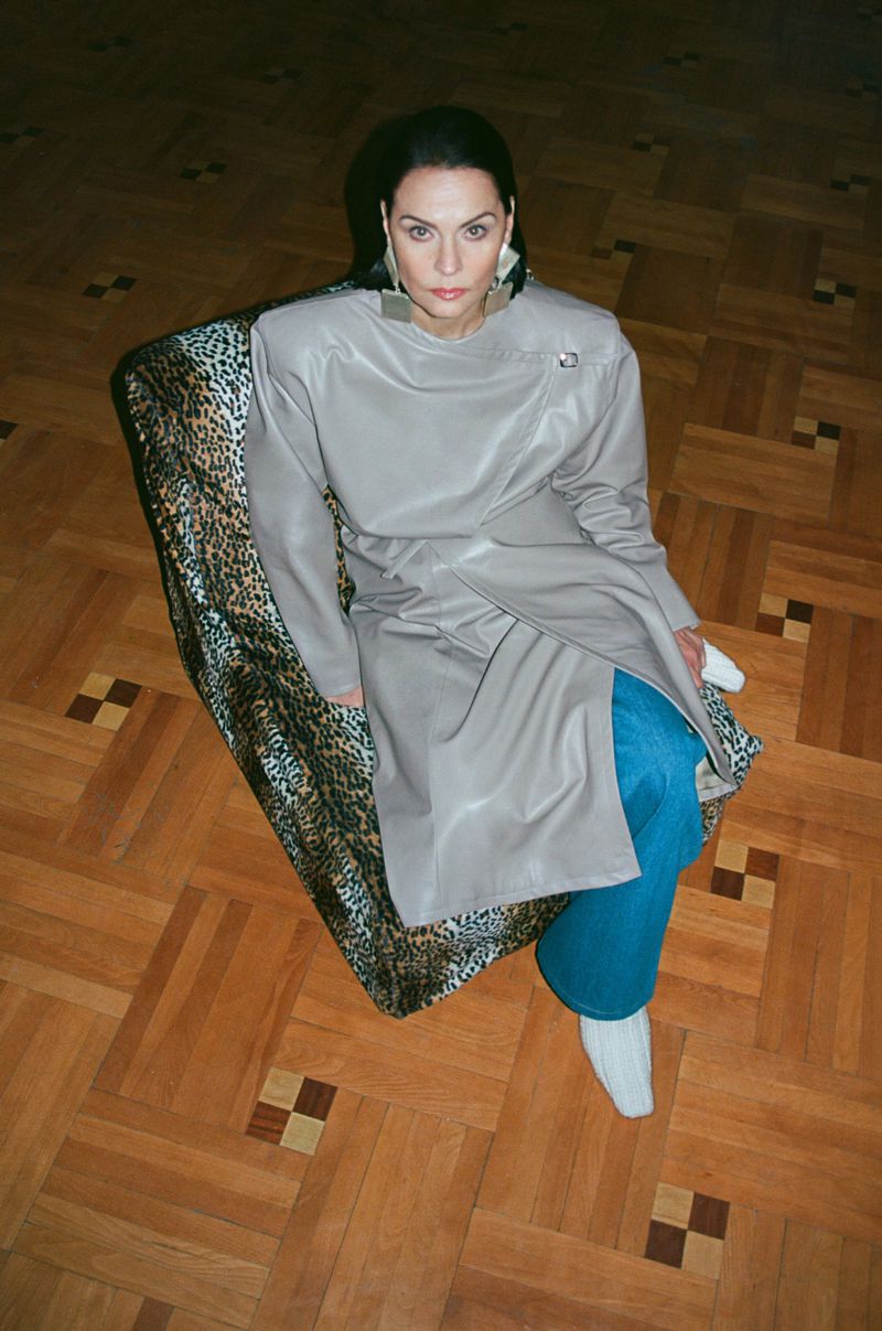 Eye-level shot of woman in gray wrap dress seated on animal-print textile, blue jeans, and white socks against wood parquet flooring.