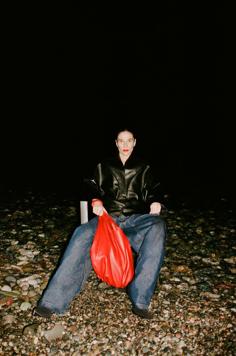 Adult female seated on a beach with a red bag, wearing jeans and a black leather jacket against a dark night background.