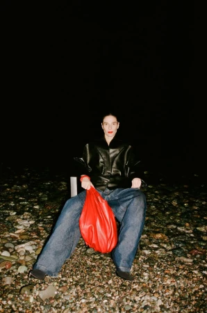 Adult female seated on a beach with a red bag, wearing jeans and a black leather jacket against a dark night background.