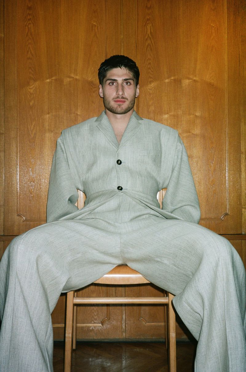 A man sits on a wooden chair against a paneled wall, wearing a light grey, oversized suit. A waist seam creates a cinched look.