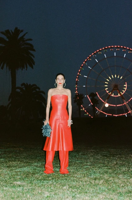 A woman in a red strapless dress and pants stands in a grassy field at night, with palm trees and a Ferris wheel in the background.
