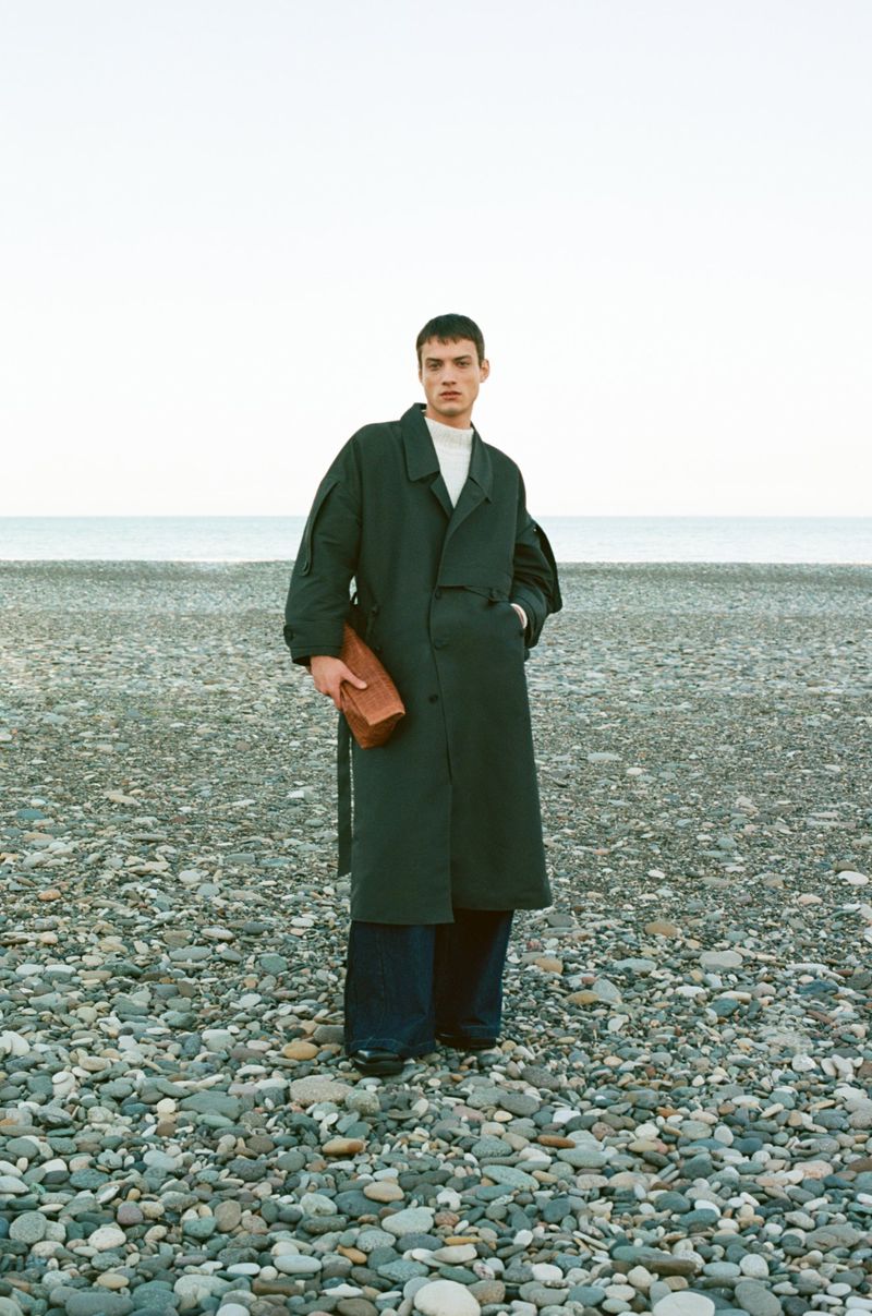 A male model standing on a pebble beach wearing a dark green coat and holding a boxy leather bag, with the ocean in the background.