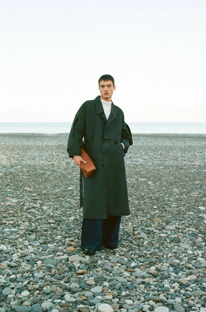 A male model standing on a pebble beach wearing a dark green coat and holding a boxy leather bag, with the ocean in the background.