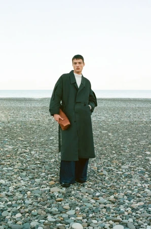 A male model standing on a pebble beach wearing a dark green coat and holding a boxy leather bag, with the ocean in the background.