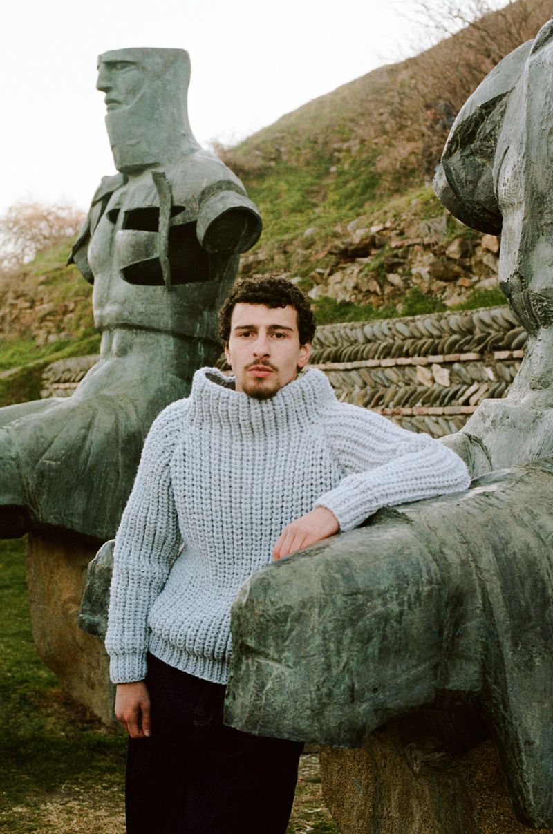 Young man poses in chunky knit sweater in front of brutalist stone sculptures and grassy hill under overcast lighting.