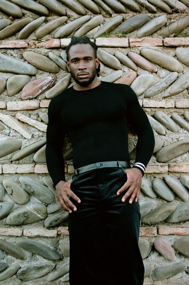 Man in black turtleneck poses against a textured wall with cobblestones and bricks, hands in pockets, fashion shoot, dark clothing.