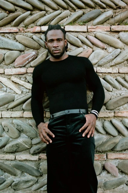 Man in black turtleneck poses against a textured wall with cobblestones and bricks, hands in pockets, fashion shoot, dark clothing.