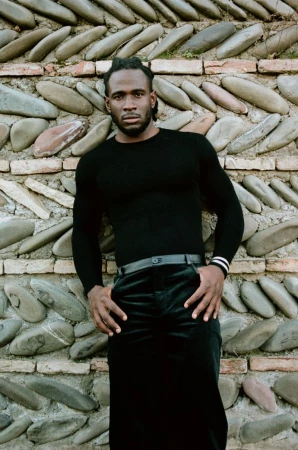 Man in black turtleneck poses against a textured wall with cobblestones and bricks, hands in pockets, fashion shoot, dark clothing.
