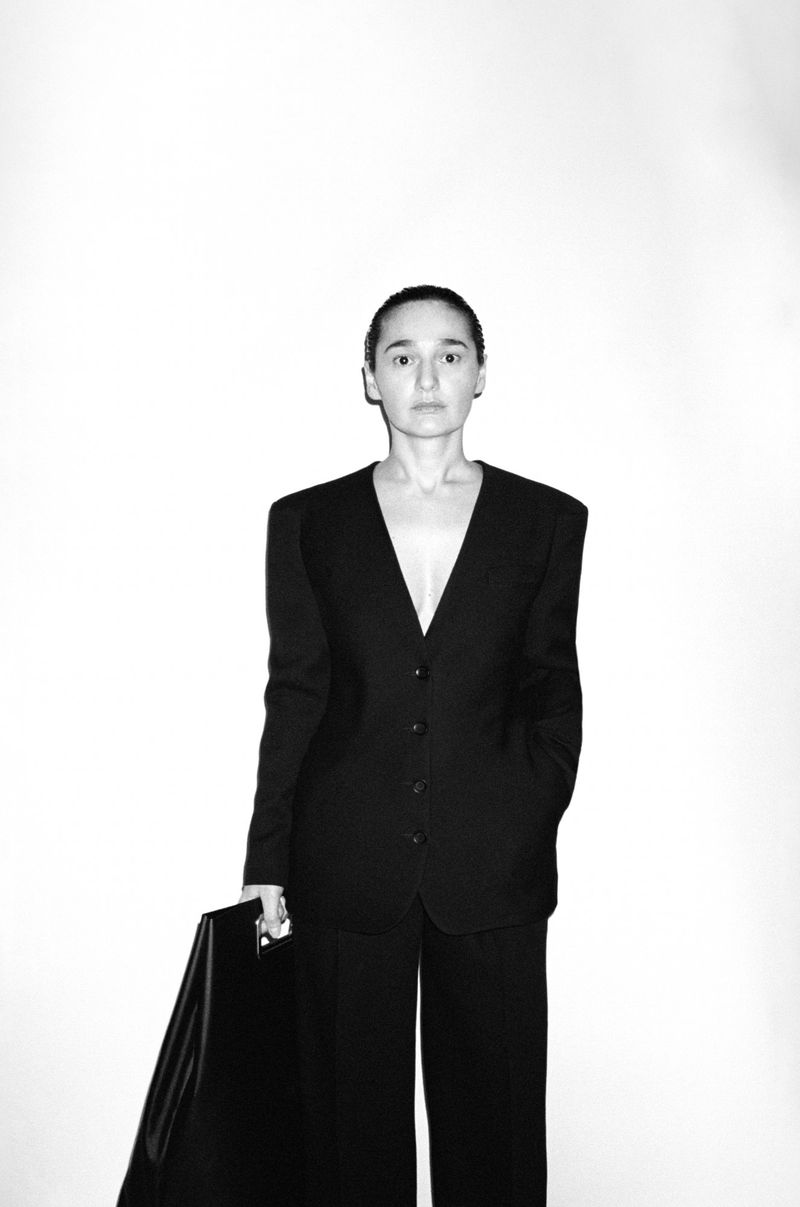 Portrait of a woman in a black suit holding a black bag against a white background in a studio, showcasing minimalist fashion.