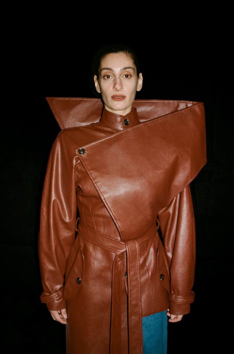 An adult female model poses in a fashionable brown leather coat dress with unique asymmetrical detailing against a black background.