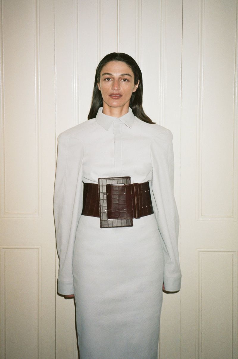 A slender adult woman with long dark hair poses indoors wearing a light gray dress and dark brown crocodile corset belt with a serious expression.