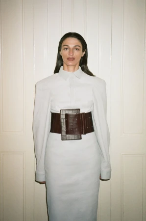 A slender adult woman with long dark hair poses indoors wearing a light gray dress and dark brown crocodile corset belt with a serious expression.