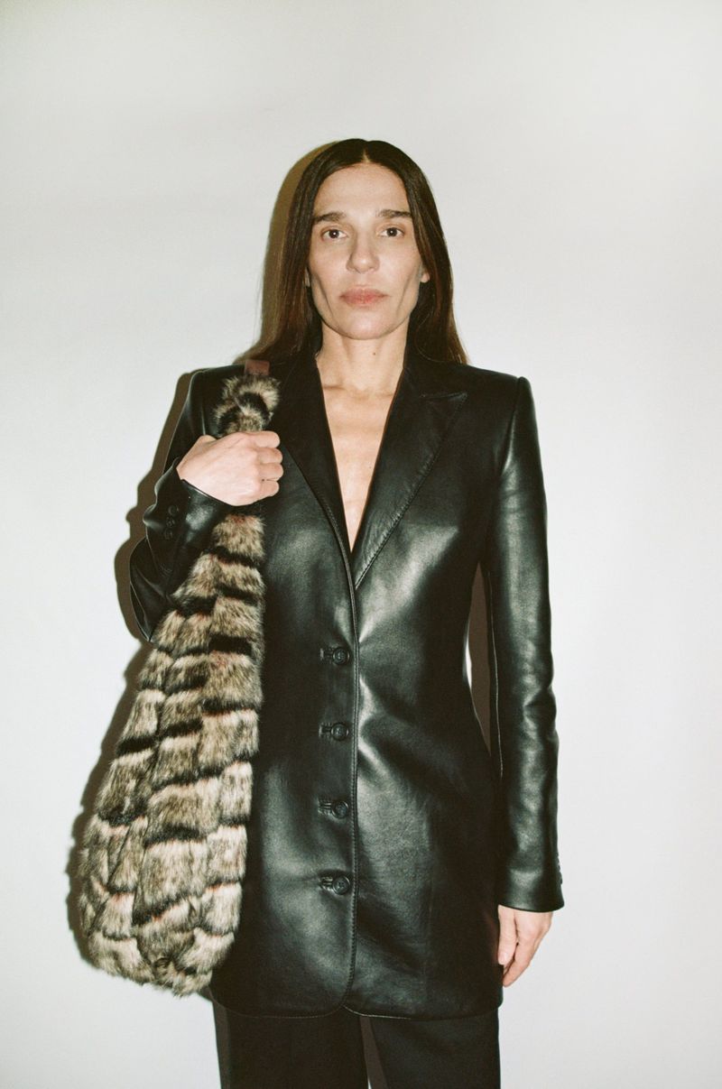 An adult woman in a black leather jacket poses indoors holding a tote bag with an animal print against a plain white background.