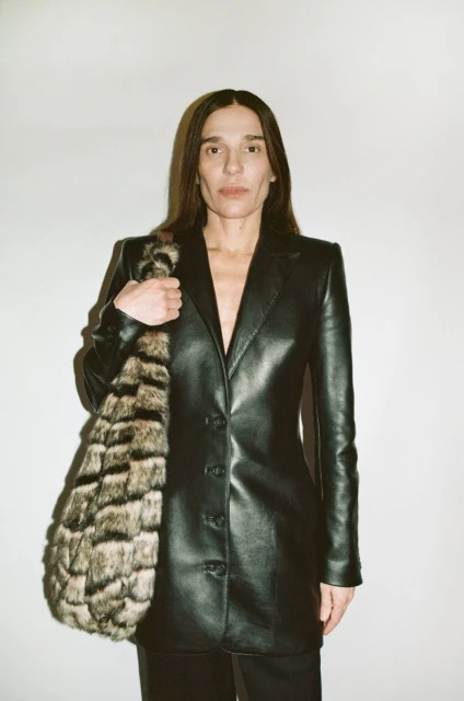 An adult woman in a black leather jacket poses indoors holding a tote bag with an animal print against a plain white background.