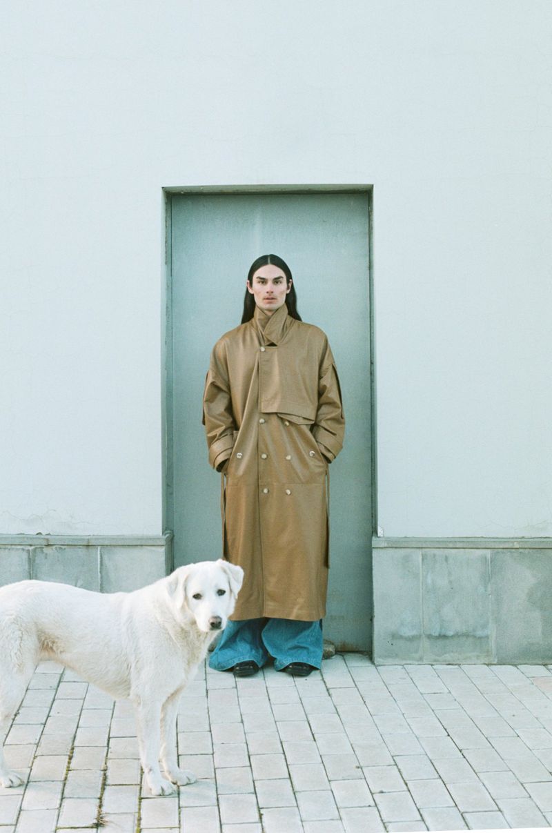 A person in a tan trench coat stands in front of a door next to a white dog, on a brick sidewalk against a light blue-gray wall.