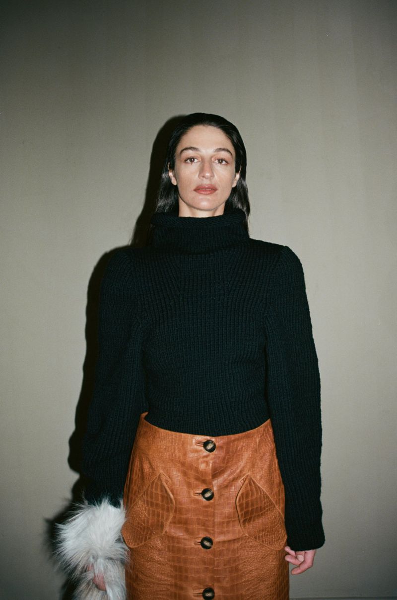 Portrait of a woman with dark hair wearing a black turtleneck sweater and brown skirt, standing against a light gray background.
