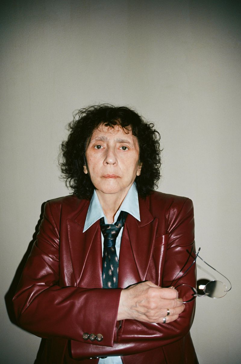 Portrait of a woman in a red leather jacket, light blue button-down shirt, and patterned tie, holding eyeglasses with a neutral expression.
