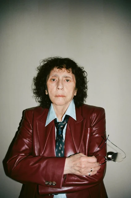 Portrait of a woman in a red leather jacket, light blue button-down shirt, and patterned tie, holding eyeglasses with a neutral expression.