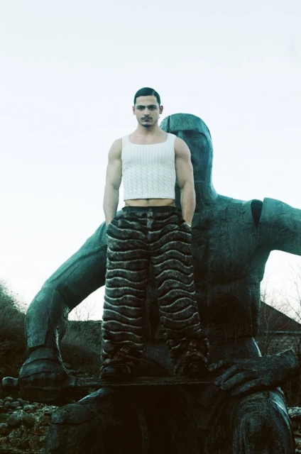 An adult male poses next to an outdoor sculpture, wearing a tank top and patterned pants in a medium shot under cloudy daylight conditions.