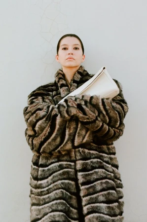 Model wearing an oversized striped faux fur coat while holding a white leather clutch bag against a pale wall.