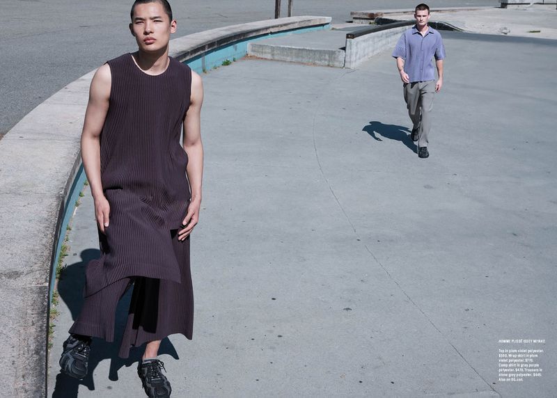 Two adult men in an outdoor setting. One man in a sleeveless outfit stands in the foreground, while the other walks in the background.