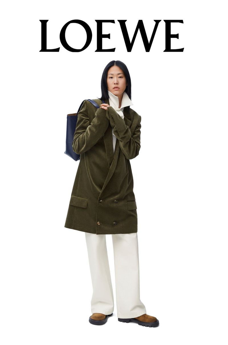 An Asian female model posing in an olive green corduroy blazer, white pants, and tan shoes. She carries a tote bag and wears a white turtleneck.