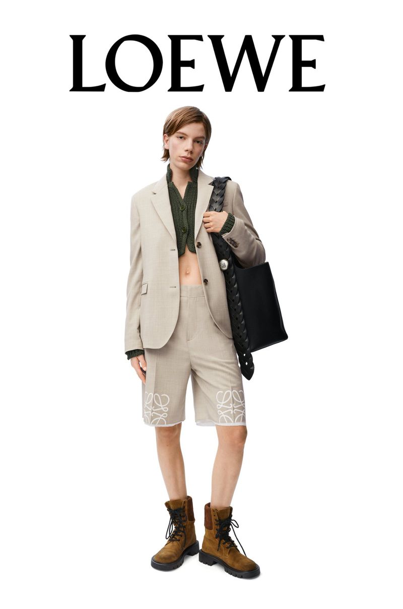 A person wearing a tan suit and brown boots stands in a studio, carrying a black bag; the LOEWE brand logo is visible above.