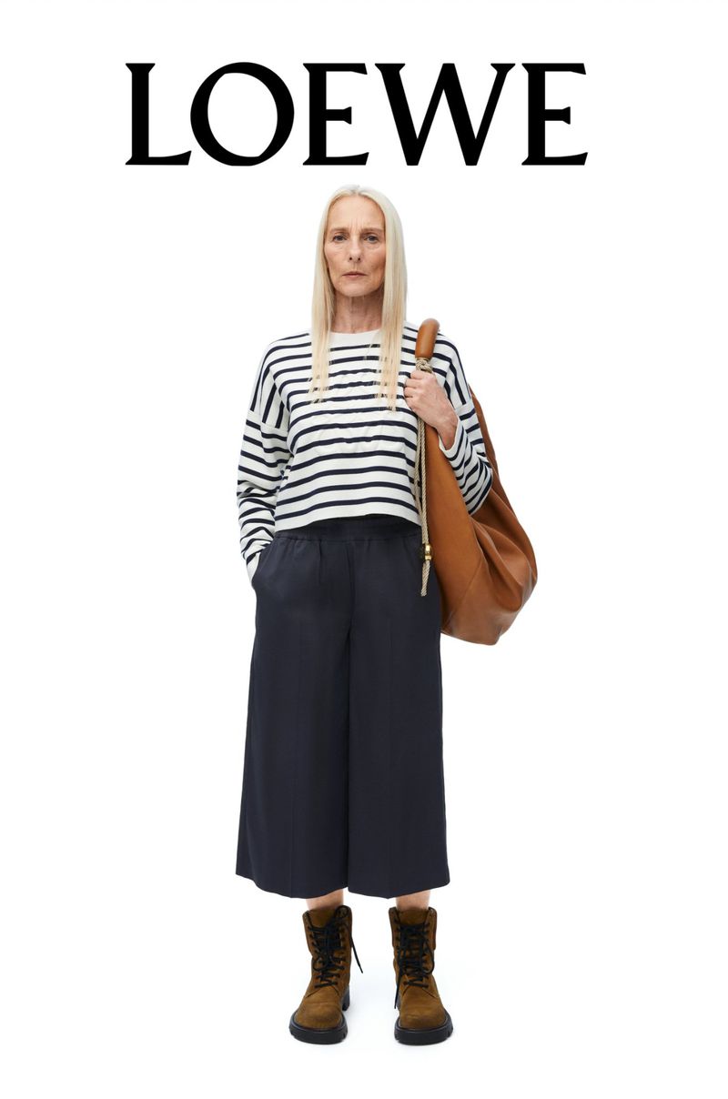 Adult woman stands in a fashion shot, wearing a striped top, culottes, and boots, posing with a brown leather hobo bag. LOEWE logo above.
