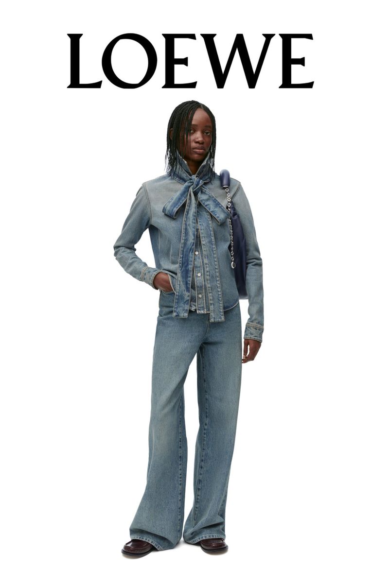 Fashion editorial shot of a woman in a blue denim outfit with a neck tie and navy handbag against a white background. Loewe text visible.
