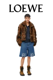 Model standing against a white seamless background wearing a brown fur jacket, dark blue buttoned shirt, denim shorts, and brown boots.
