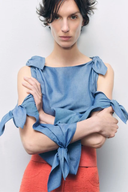 Adult woman in a light-blue bow-tied top and salmon-colored bottoms poses against a white background in a fashion-style portrait.