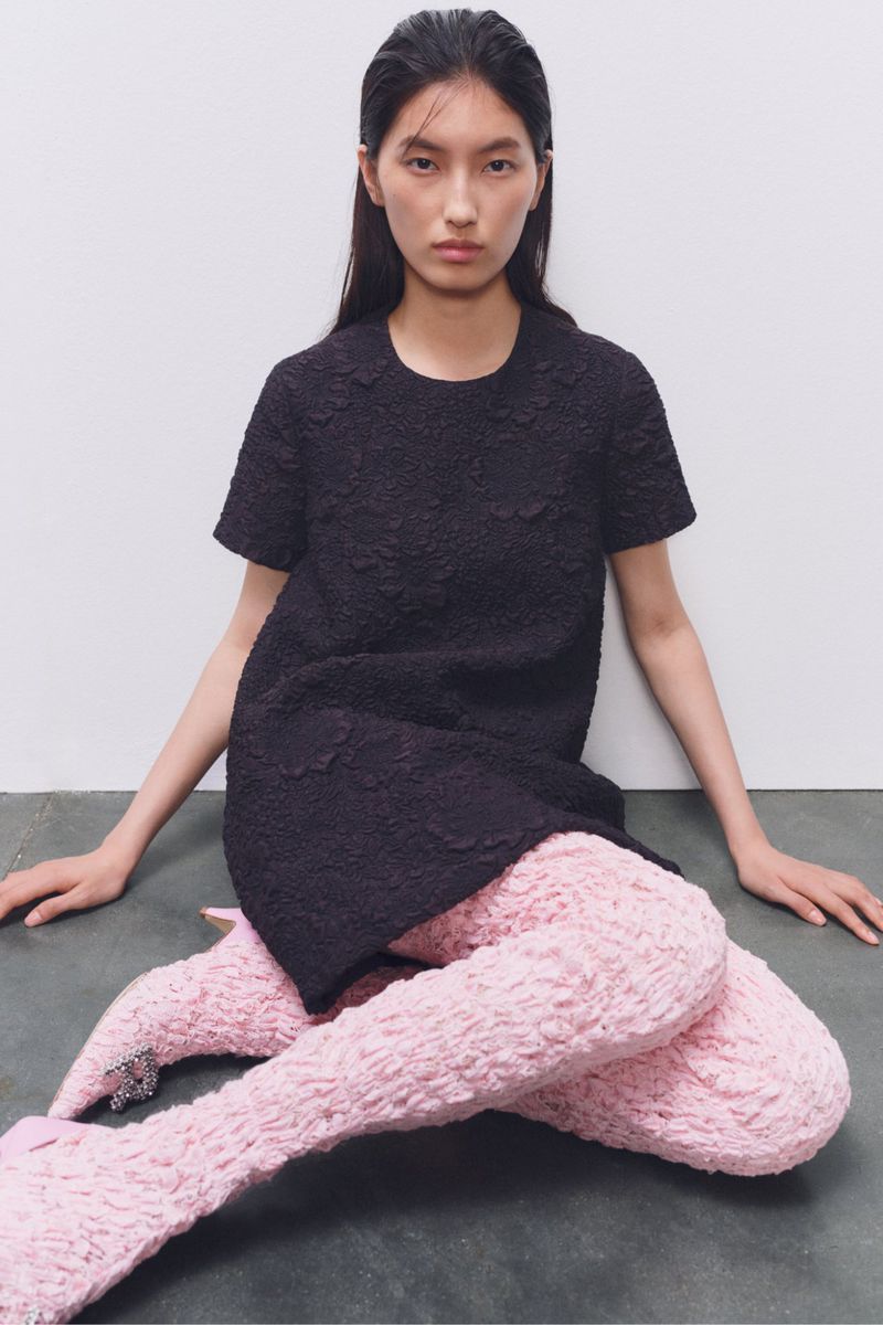 Fashion photo: Woman in a black textured mini dress sits against a gray background, wearing light pink textured tights and bejeweled shoes.
