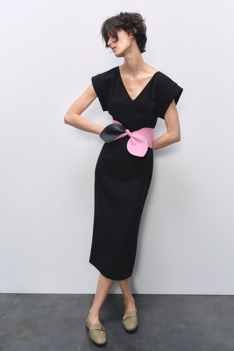 A slender woman poses in a sleek black midi dress with a statement pink and black belt against a plain grey backdrop.