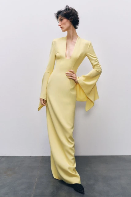 A slender woman models a pale yellow gown with flared sleeves, set against a minimalist backdrop in a fashion studio.